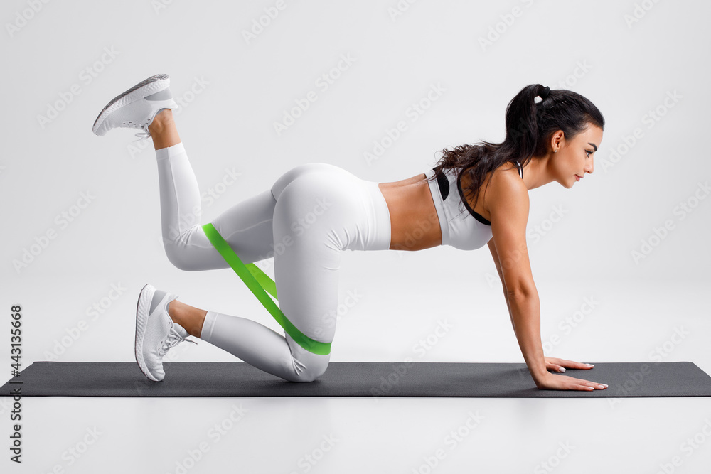 Fitness woman doing kickback exercise for glutes with resistance band on gray background. Athletic girl working out donkey kicks. - obrazy, fototapety, plakaty 