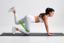 Fitness Woman Doing Kickback Exercise For Glutes With Resistance Band On Gray Background. Athletic Girl Working Out Donkey Kicks.