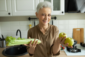 Smiling middle-aged woman posing in home kitchen and showing celery and apples for healthy breakfast. Proper nutrition and diet concept. Kitchen interior on blurred background