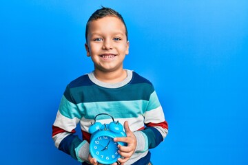 Wall Mural - Little kid boy holding alarm clock looking positive and happy standing and smiling with a confident smile showing teeth