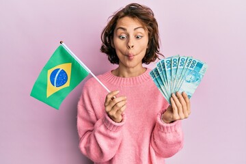 Sticker - Young brunette woman holding brazil flag and real banknotes making fish face with mouth and squinting eyes, crazy and comical.