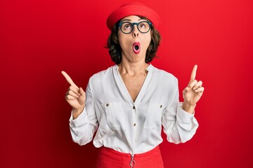 Poster - Young brunette woman wearing stewardess style and glasses amazed and surprised looking up and pointing with fingers and raised arms.