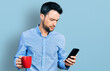 © Krakenimages.com - Hispanic man with beard using smartphone and drinking a cup of coffee skeptic and nervous, frowning upset because of problem. negative person.