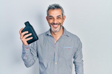 Wall Mural - Handsome middle age man with grey hair holding motor oil bottle looking positive and happy standing and smiling with a confident smile showing teeth