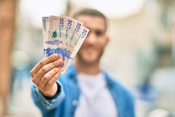 Sticker - Young caucasian man smiling happy holding colombian pesos at the city.