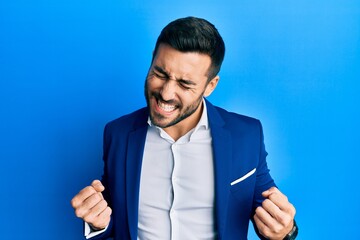 Wall Mural - Young hispanic businessman wearing business jacket excited for success with arms raised and eyes closed celebrating victory smiling. winner concept.