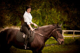Fototapeta Konie - Young woman riding a black horse in the paddock.