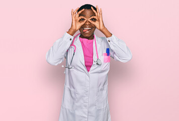 Canvas Print - Young african american woman wearing doctor uniform and stethoscope doing ok gesture like binoculars sticking tongue out, eyes looking through fingers. crazy expression.