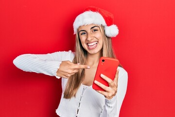 Poster - Beautiful hispanic woman wearing christmas hat holding smartphone smiling happy pointing with hand and finger