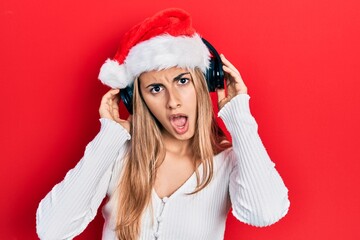 Poster - Beautiful hispanic woman wearing christmas hat and headphones in shock face, looking skeptical and sarcastic, surprised with open mouth