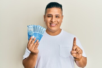 Canvas Print - Young latin man holding 10000 chilean pesos smiling with an idea or question pointing finger with happy face, number one