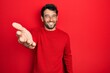 Handsome man with beard wearing casual red sweater smiling cheerful offering palm hand giving assistance and acceptance.