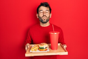 Sticker - Handsome man with beard eating a tasty classic burger with fries and soda looking at the camera blowing a kiss being lovely and sexy. love expression.