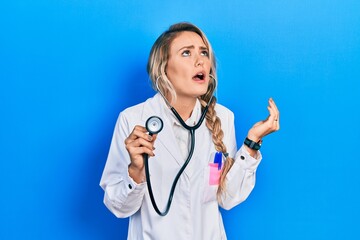 Sticker - Beautiful young blonde doctor woman holding stethoscope crazy and mad shouting and yelling with aggressive expression and arms raised. frustration concept.