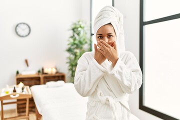 Canvas Print - Young brunette woman wearing towel and bathrobe standing at beauty center shocked covering mouth with hands for mistake. secret concept.