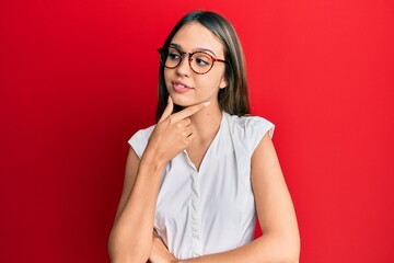 Wall Mural - Young brunette woman wearing casual clothes and glasses with hand on chin thinking about question, pensive expression. smiling with thoughtful face. doubt concept.