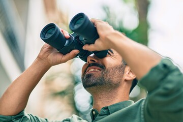 Canvas Print - Young hispanic man smiling happy looking for new opportunity using binoculars at the city.