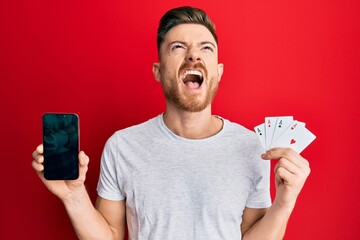 Wall Mural - Young redhead man holding smartphone and casino card angry and mad screaming frustrated and furious, shouting with anger looking up.