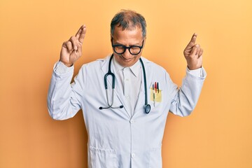 Wall Mural - Middle age indian man wearing doctor coat and stethoscope gesturing finger crossed smiling with hope and eyes closed. luck and superstitious concept.