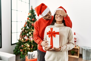 Young hispanic man surprisng his girlfriend with christmas gift at home.