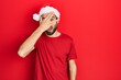© Krakenimages.com - Young hispanic man wearing christmas hat peeking in shock covering face and eyes with hand, looking through fingers with embarrassed expression.