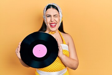 Sticker - Young hispanic woman wearing pin up style holding vinyl disc winking looking at the camera with sexy expression, cheerful and happy face.