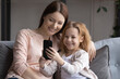 © fizkes - Smiling junior school age girl sit on sofa with young adult mother use smartphone take selfie for social media watch cartoon. Happy preteen child daughter and millennial mom make videocall at cell app