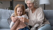 © fizkes - Granny teaching to knit. Positive elderly grandma embrace little girl grandchild sitting on couch holding needles and yarn show pattern sample. Skilled grandmother enjoy needlework with small grandkid