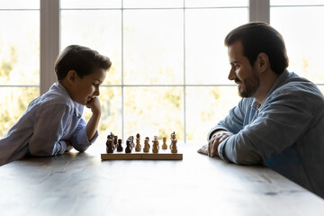 Focused thoughtful clever son and dad playing chess, sitting at table with chessboard, thinking over game strategy, planning next move, training logical and tactic skills. Family hobby concept