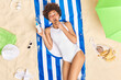 Overhead shot of shocked ethnic female model holds sunscreen lotion has burned skin after long time sunbathing wears white bikini poses on white sand surrounded by beach accessories. Sunburn