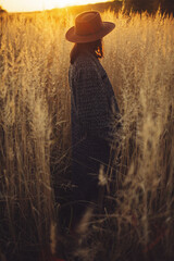 Wall Mural - Stylish woman in hat standing in grass in warm sunset light in autumn field. Tranquil atmospheric moment. Fashionable young female in retro clothes enjoying evening sunshine in countryside