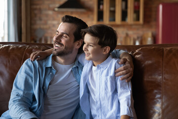 Wall Mural - Happy preschooler son and daddy sitting together on sofa, talking and hugging, looking away, thinking, planning future. Father and kid enjoying being friends, spending leisure time together at home