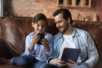 Wall Mural - Happy gen Z boy kid and daddy using gadgets at home, playing, compete in online game, holding digital tablet and mobile phone, browsing internet, using learning app, making video call. Family activity