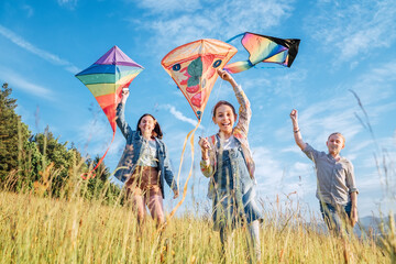 Wall Mural - Smiling gils and brother boy running with flying colorful kites on the high grass meadow in the mountain fields. Happy childhood moments or outdoor time spending concept image.