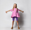 Cheerful laughing blonde 6-7 years old. a girl with a girly hairstyle in a pink sweater with a peplum and legends stands with her arms outstretched on a gray background