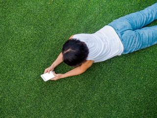 Beautiful happy Asian woman black short hair in white sleeveless shirt and blue jeans looking at smartphone with smiling while lying on green artificial grass, top view.