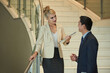 © DragonImages - Beautiful elegant young businesswoman with documents folder talking to colleague when standing on staircase in lobby of business center