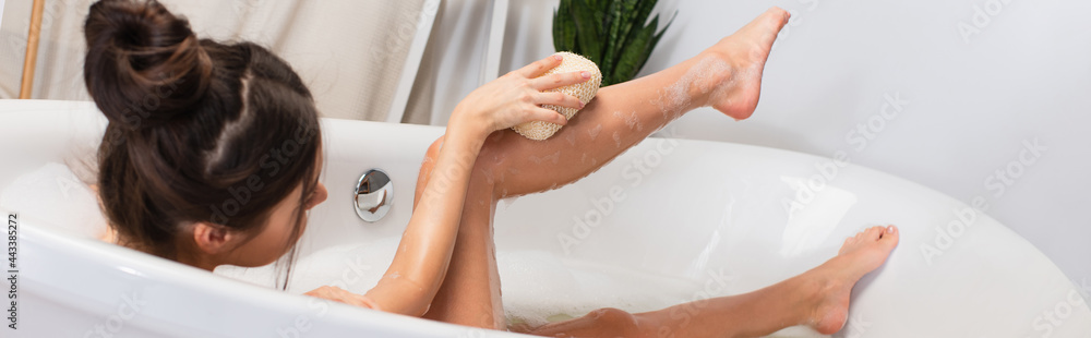 young woman with hair bun taking bath with loofah in bathtub, banner - obrazy, fototapety, plakaty 
