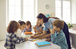 © Studio Romantic - Happy teacher and children reading book together. Group of school kids gather around table in classroom to listen to interesting story told by teacher. Elementary students learning new things in class