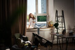 © olezzo - Beautiful happy young woman using smartphone sitting on windowsill in modern apartment with laptop on the table