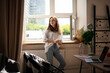 © olezzo - Young beautiful brunette woman drinking morning coffee while standing by the window in the apartment near table with laptop