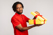 Disappointed man with dreadlocks in red casual T-shirt, holding unpacked gift box, has frustrated dissatisfied facial expression, upset with present. Indoor studio shot isolated on gray background.