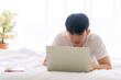 © kaew6566 - New normal, social distance or technology concept. Freelancer young man casual using laptop tele working online or video conference on white bed beside windows while after awake in morning at home.