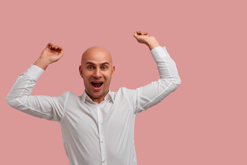 Good news. Ecstatic overjoyed bald man with bristle, clenches fists in triumph, happy with sales, dressed in white shirt. Isolated over pink background witn empty space for your advertising.