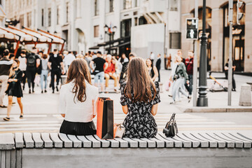 Two girls sitting on bench, summer day, view from behind, blurred pedestrians. Regular people out in public places