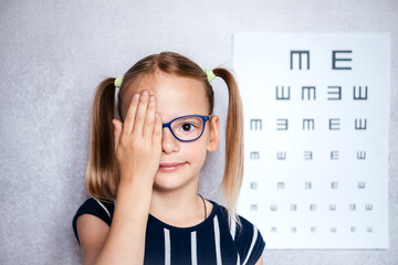 Wall Mural - Little girl wearing eyeglasses taking eyesight test before school with blurry eye chart at the background