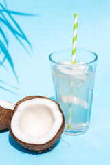 Wall Mural - A glass of fresh organic coconut water, milk on a blue pastel background and a ripe half of a coconut next to under palm leaves. Refreshing vegetarian drinks