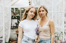 Mom And Daughter Teenage Girl In Jeans And White T-shirts Stand Near White Veranda In The Summer Garden Decorated With Vintage Details And Bouquets Of Wildflowers Lupines, Concept Of Parents And Teens