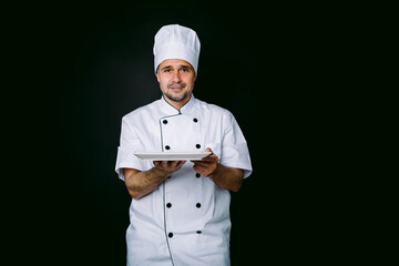 Chef cook wearing cooking jacket and hat, holding a plate, on black background