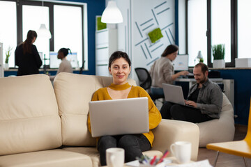 Poster - Businesswoman sitting on couch holding laptop, smiling at camera while diverse colleagues working in background. Multiethnic coworkers talking about start up financial company in modern business